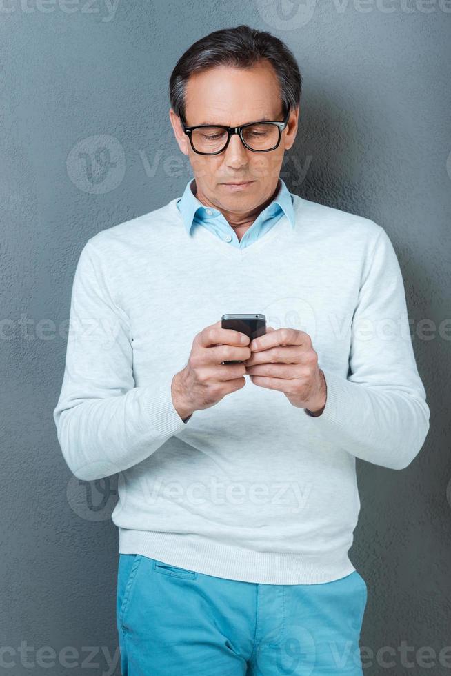 Using his phone for being in touch. Confident mature man holding mobile phone while standing against grey background photo