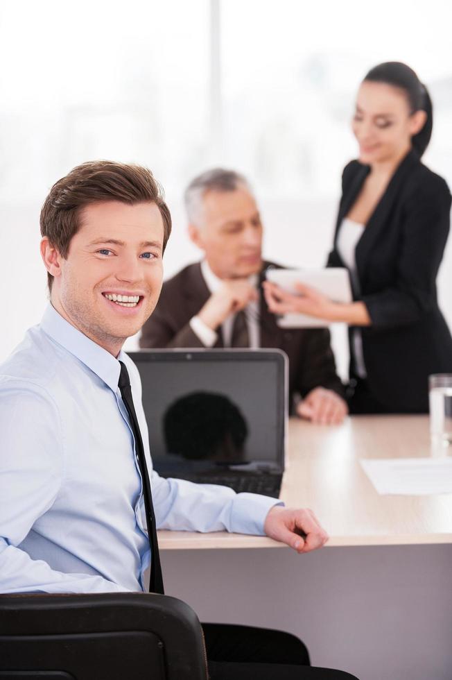 I love my job. Handsome young man in formalwear looking over shoulder and smiling while sitting at the table with two other business people talking on background photo