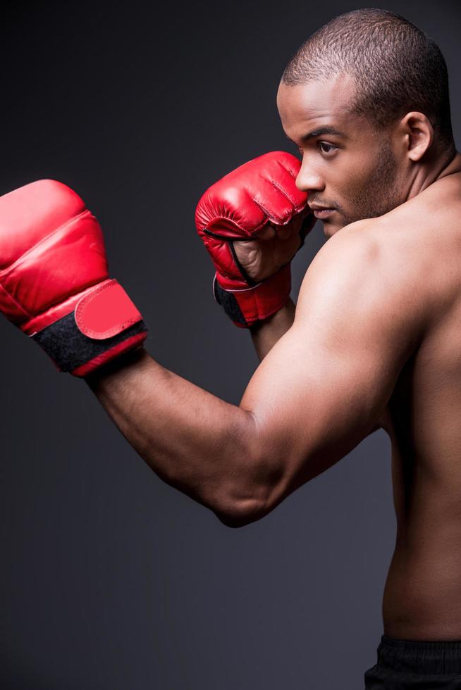 boxeador confiado. vista lateral de un joven africano sin camisa con guantes de boxeo haciendo ejercicio mientras se enfrenta a un fondo gris foto