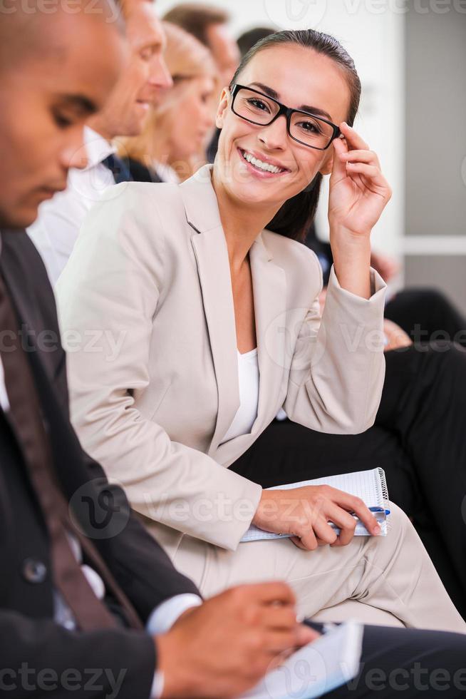 Confident business expert. Side view of business people sitting in a row and writing something in their note pads while confident young woman adjusting her eyeglasses and smiling photo