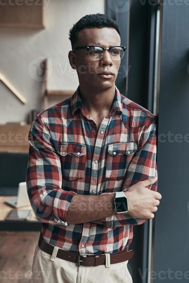 Hard decision to make... Serious young man keeping arms crossed and looking away while standing in the creative working space photo
