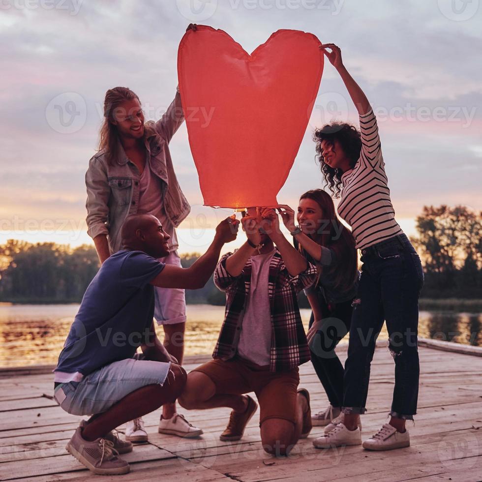 linterna en forma de corazón. toda la longitud de los jóvenes en ropa casual iluminando la linterna del cielo mientras están de pie en el muelle foto