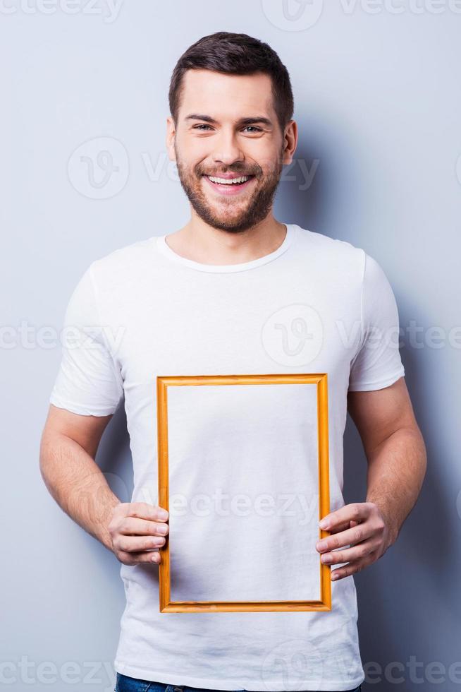Copy space in frame. Handsome young man in T-shirt holding a picture frame while standing against grey background photo