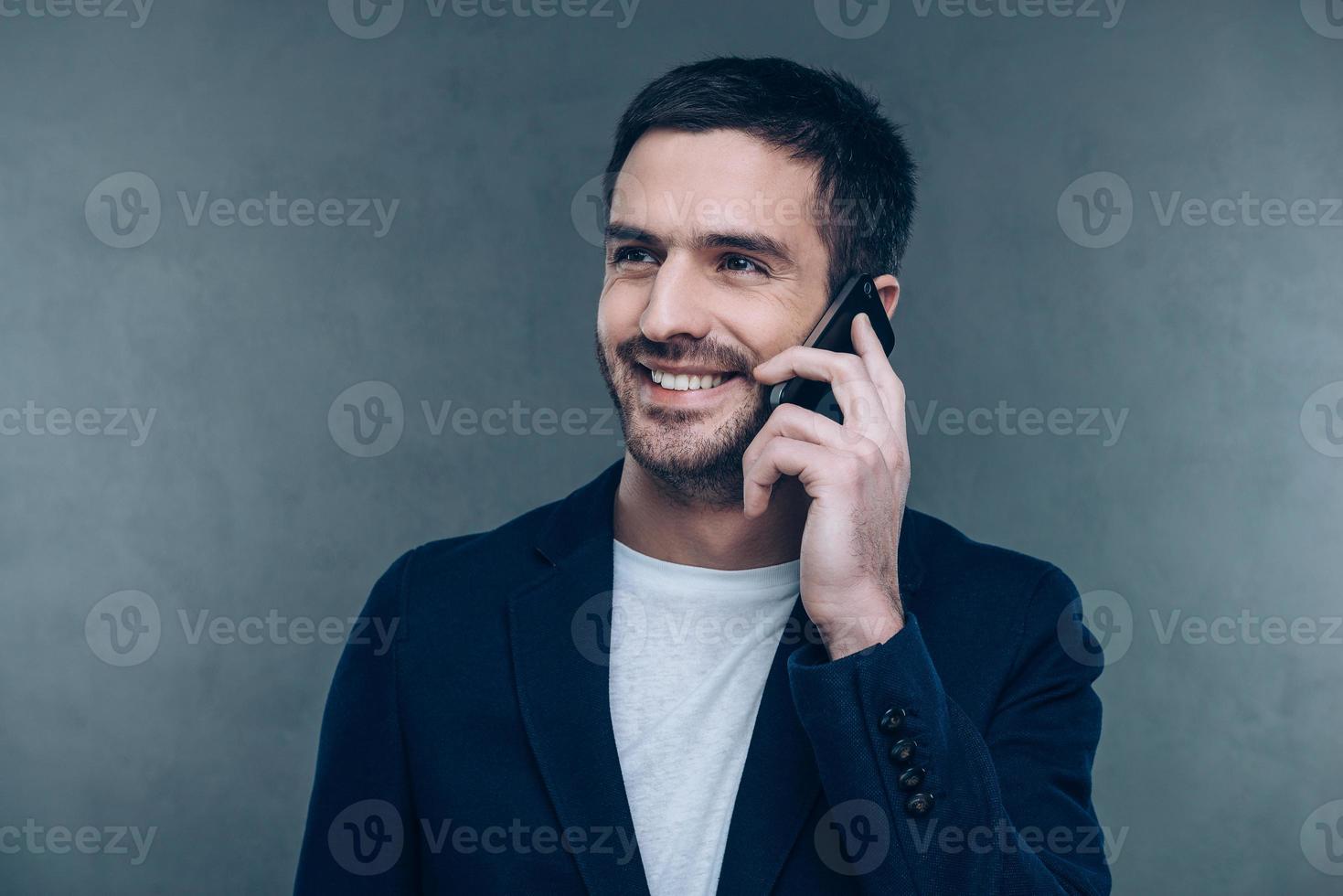 Good business talk. Cheerful young man talking on the mobile phone and smiling while standing against grey background photo