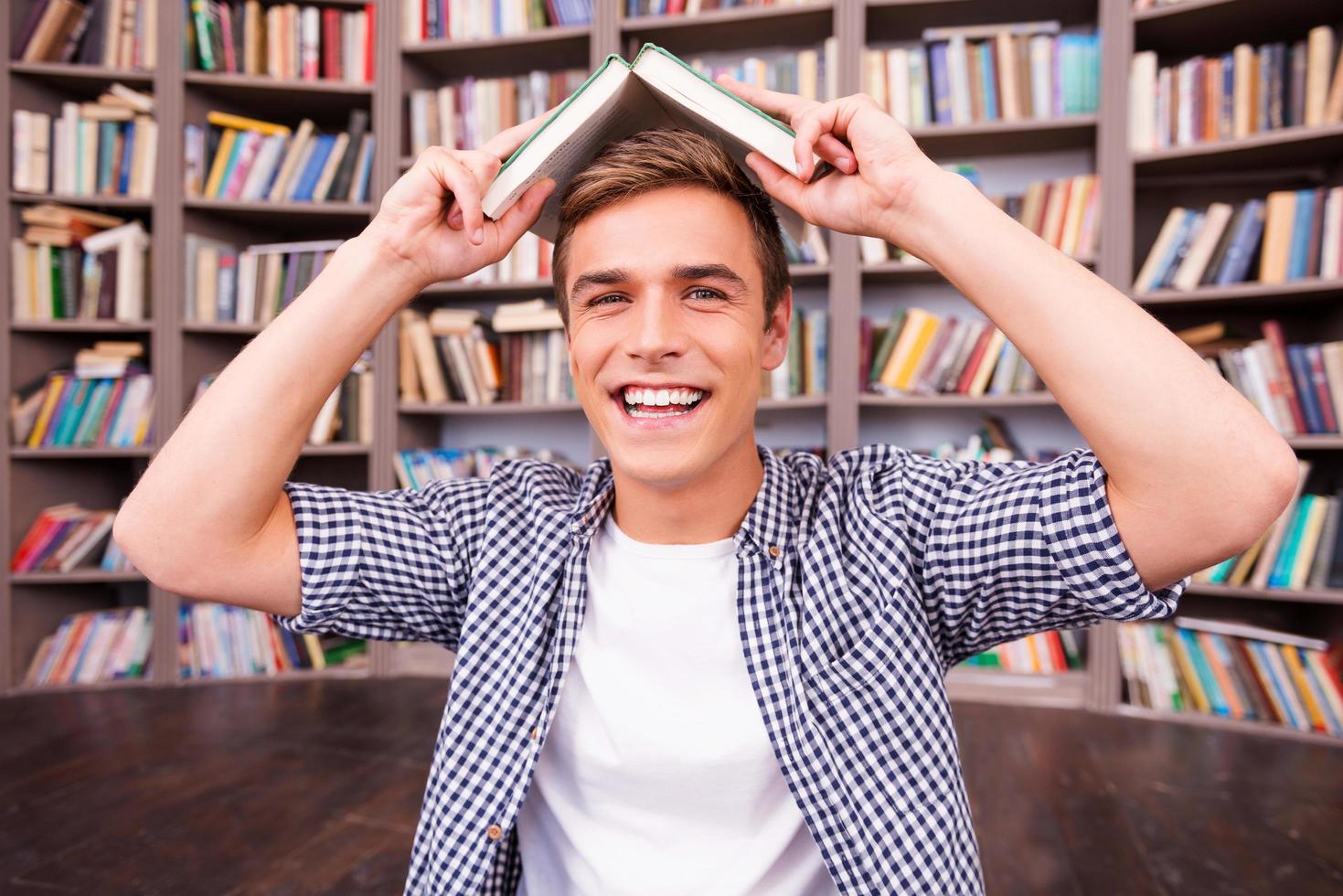 Feeling playful. Cheerful young man carrying book on head and smiling while sitting against bookshelf photo