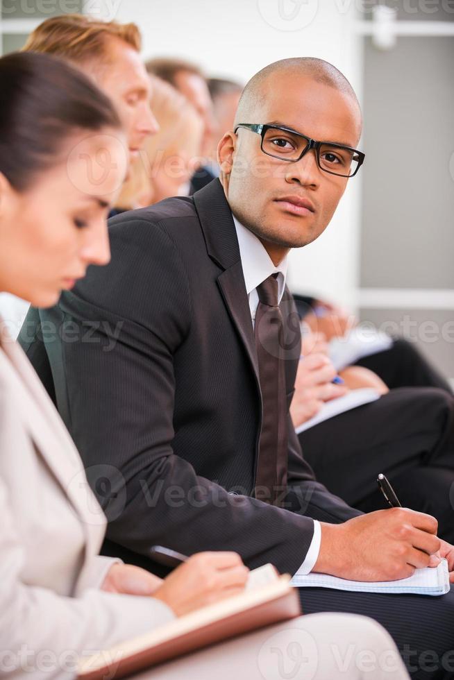 Businessman at the conference. Side view of business people sitting in a row and writing something in their note pads while confident African man in eyewear looking at camera photo