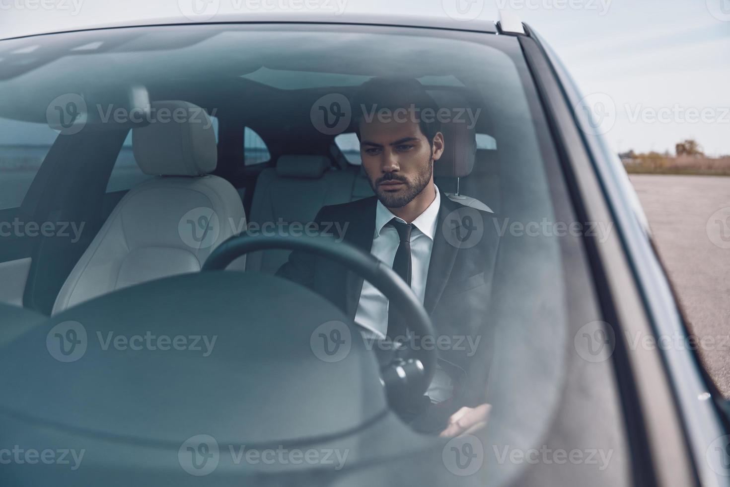 Hurrying to get things done. Handsome young man in full suit looking straight while driving a car photo