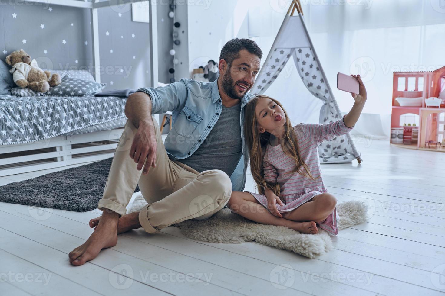 Funny selfie. Young father and his little daughter taking selfie while sitting on the floor in bedroom with the tent in the background photo