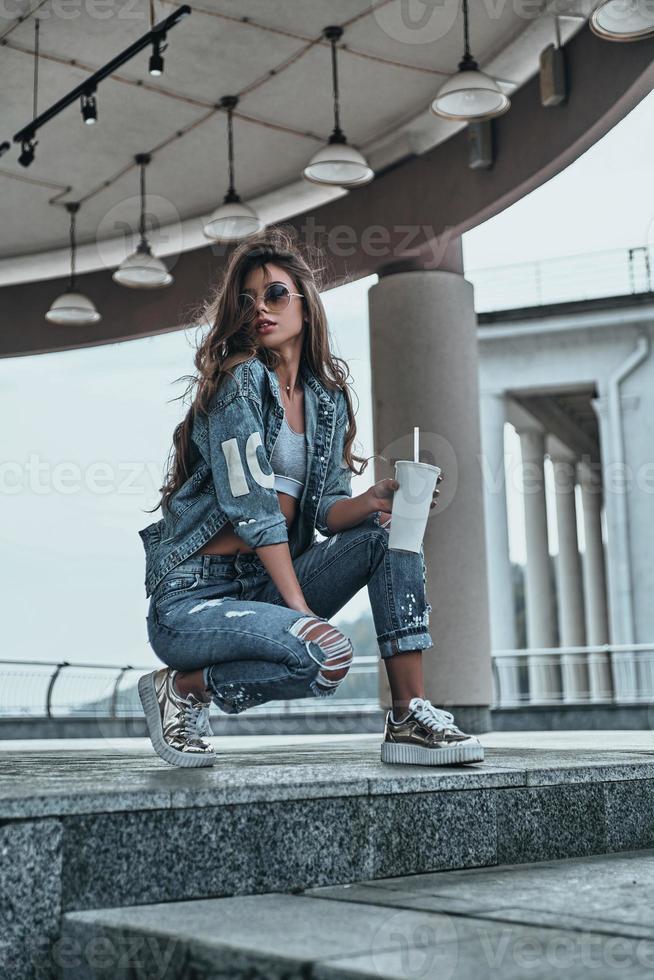 Great look. Beautiful young woman in jeans wear looking away and holding a disposable cup while spending time outdoors photo