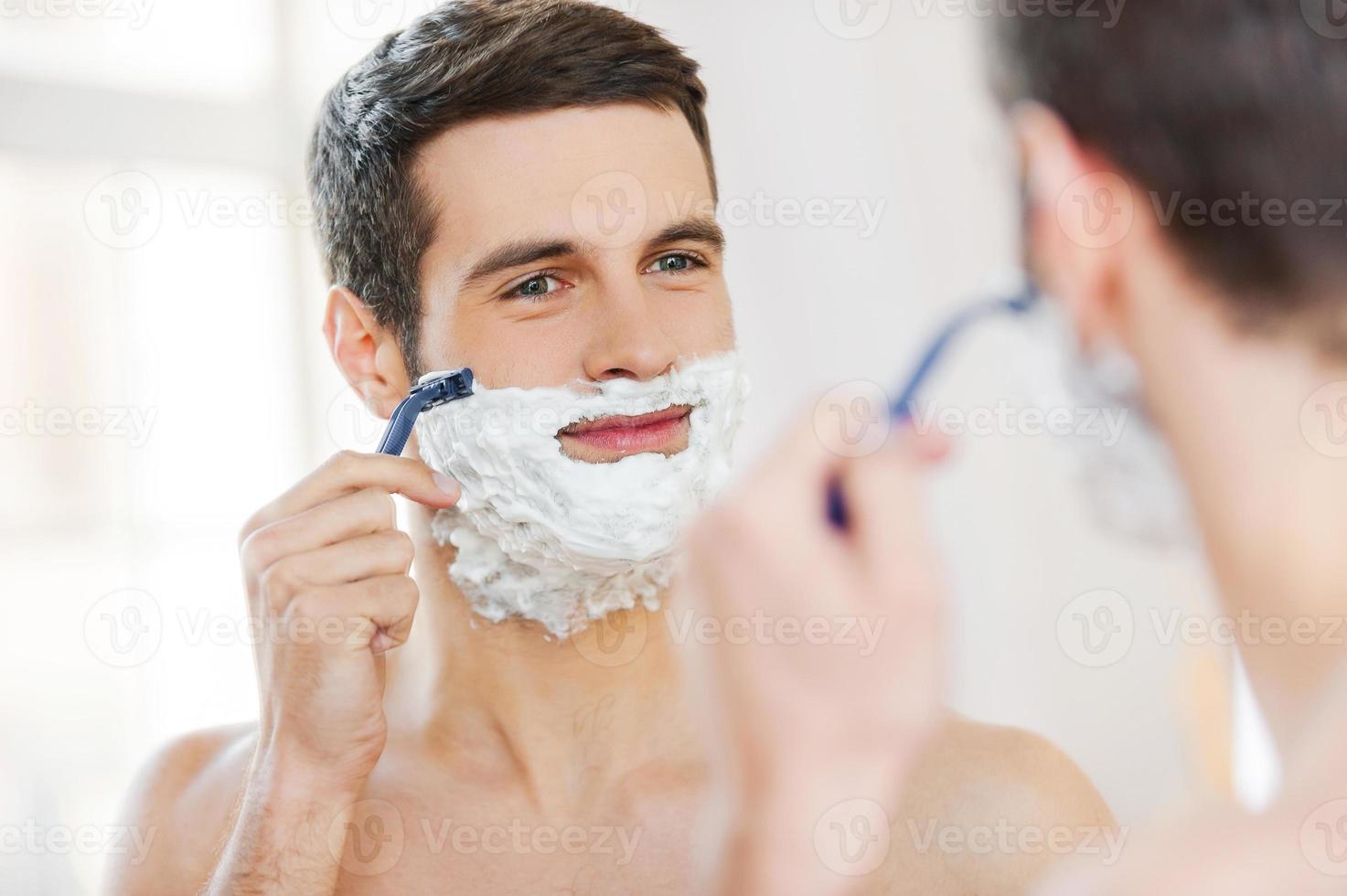Morning routine. Handsome shirtless young man shaving his face and smiling while standing in front of the mirror photo