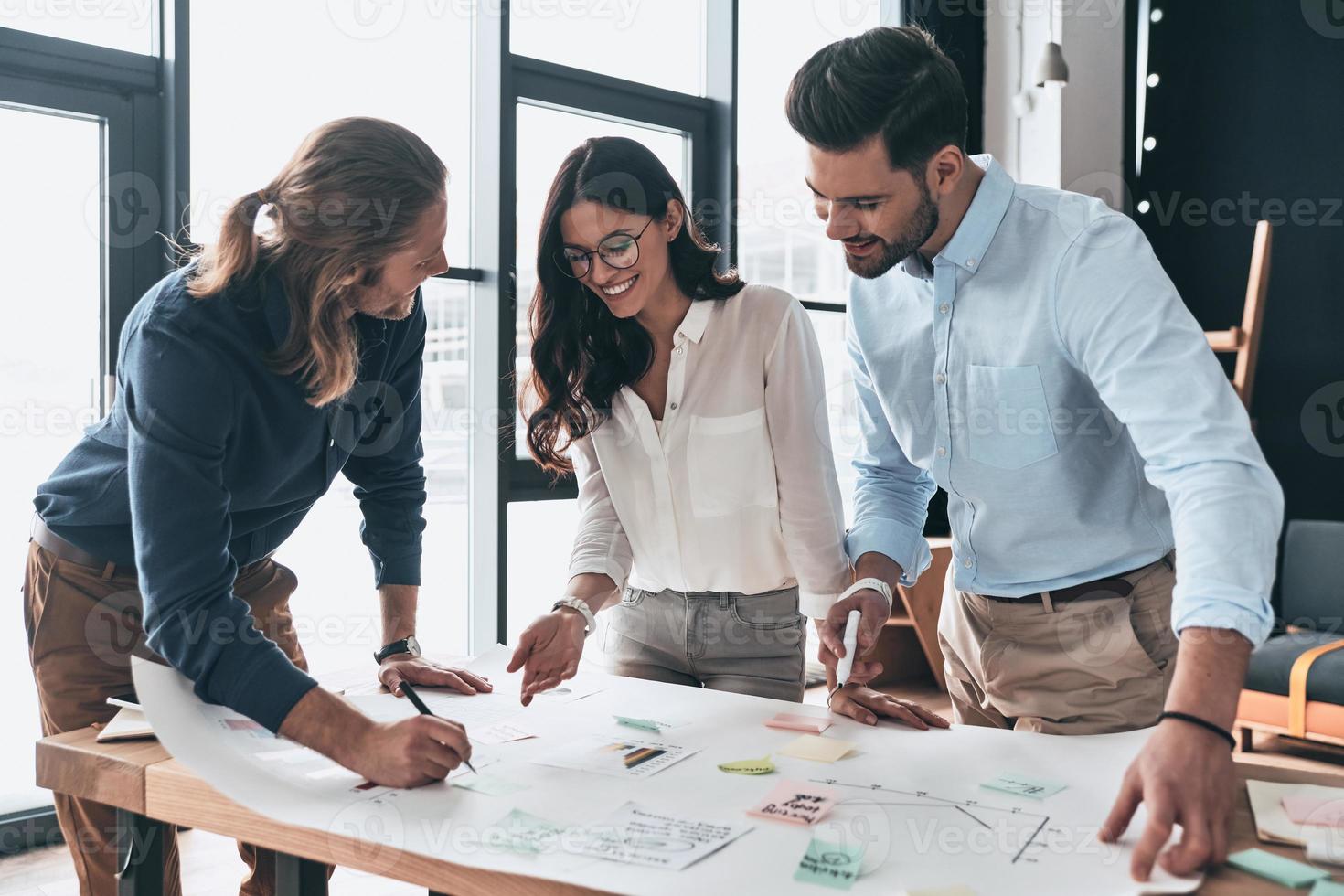 Another project. Group of young confident business people working together and smiling while man writing on blueprint in the office photo