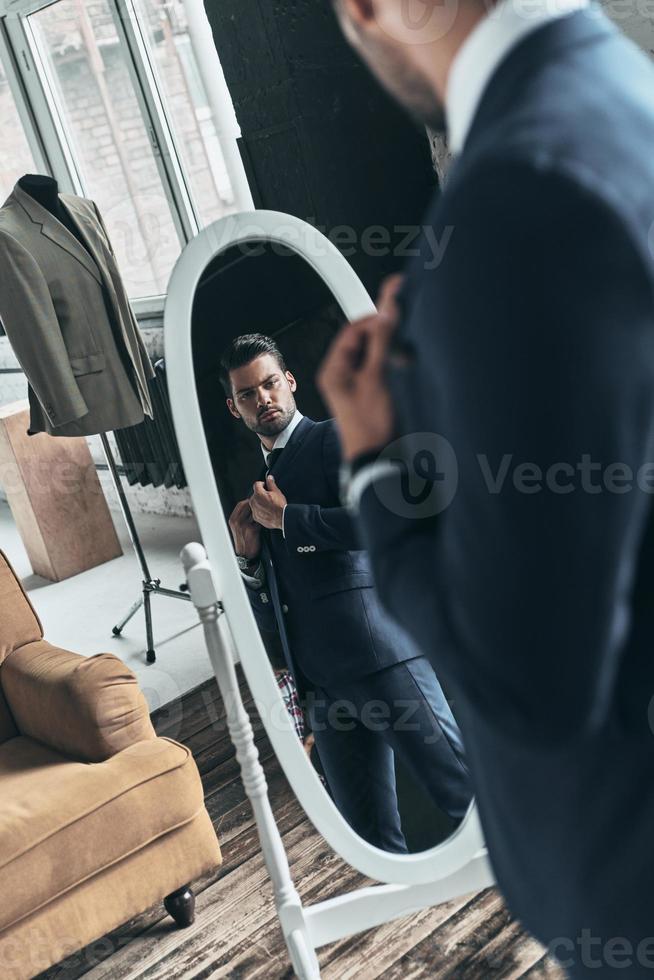 buscando simplemente perfecto. reflejo de un apuesto joven con traje completo ajustando su chaqueta mientras está de pie frente al espejo en el interior foto