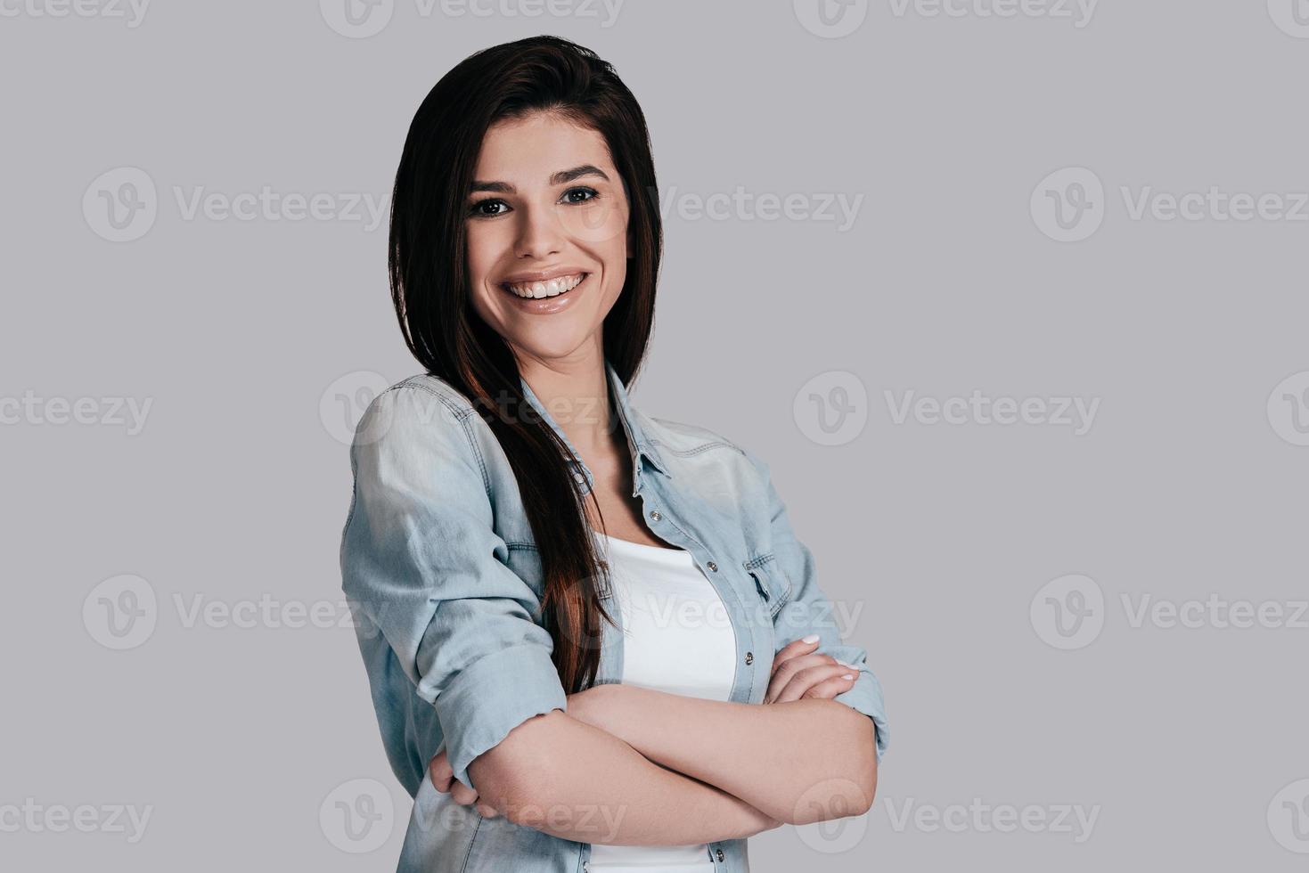 joven y hermoso. hermosa mujer joven en jeans usa manteniendo los brazos cruzados y mirando a la cámara con una sonrisa mientras está de pie contra el fondo gris foto