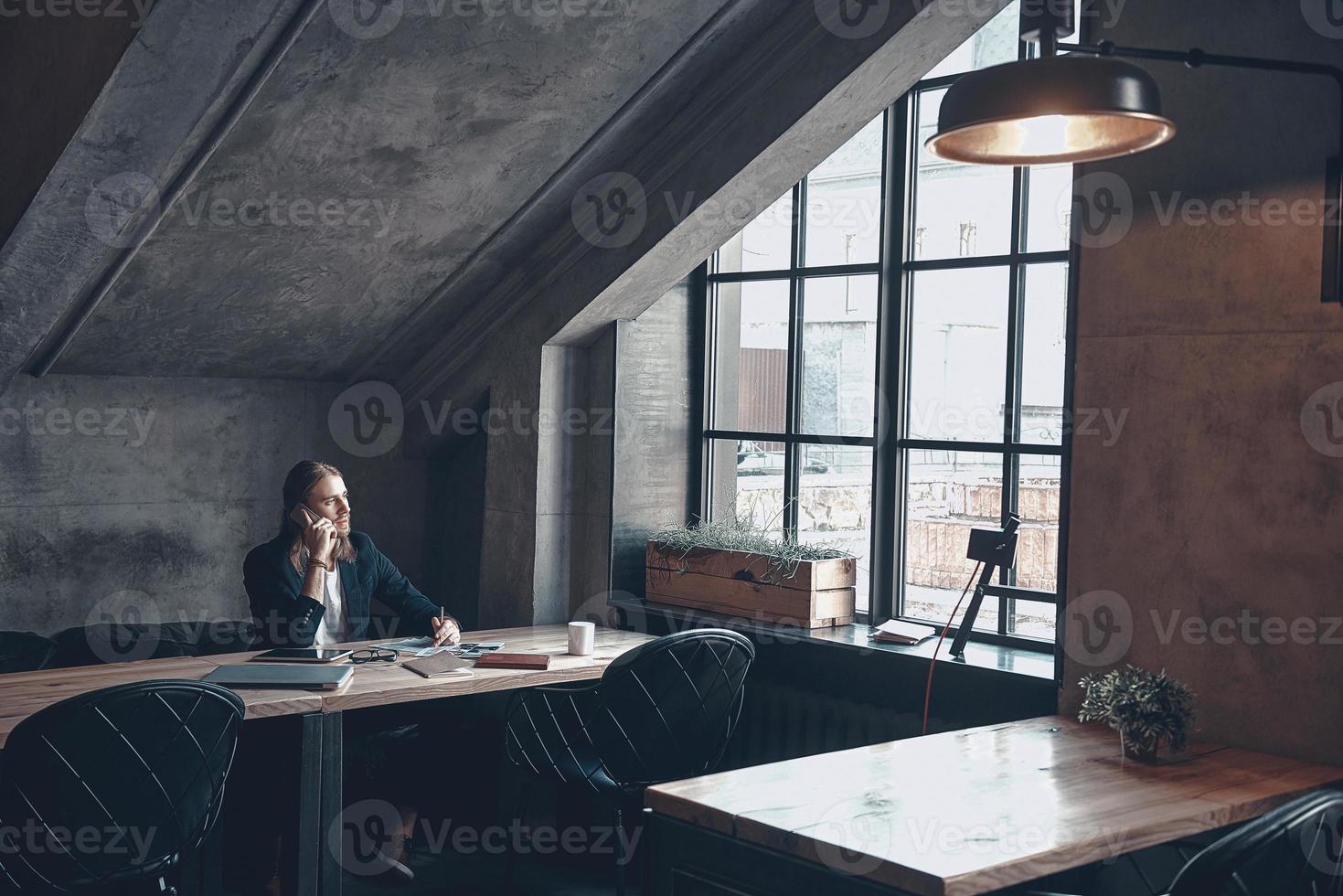 Listening to clients requirements.  Serious young man in smart casual wear talking on the smart phone and writing something down while sitting at his desk in the office photo