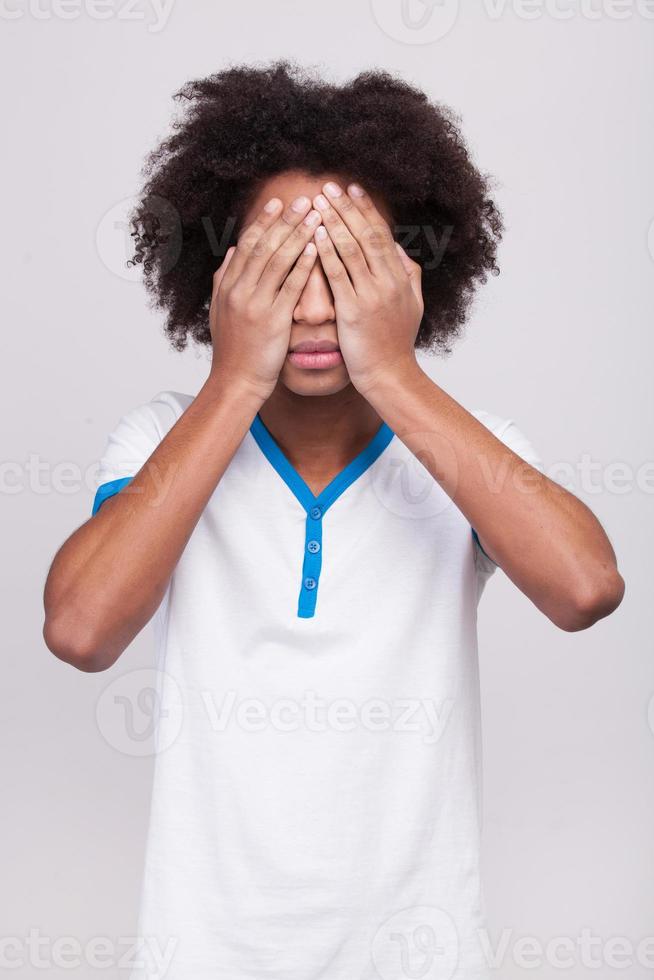 I see nothing. Teenage African boy covering eyes by hands while standing isolated on grey background photo