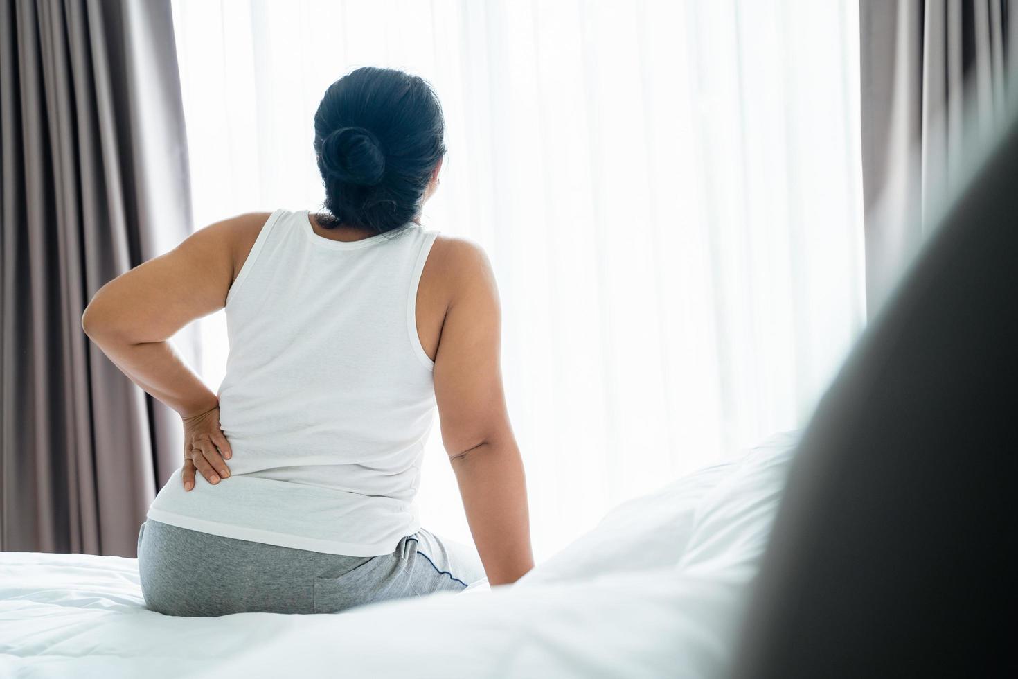 Adult Asian woman is sitting on the bed and holding her lower back suffering from injured back. Health care and back pain concept. photo