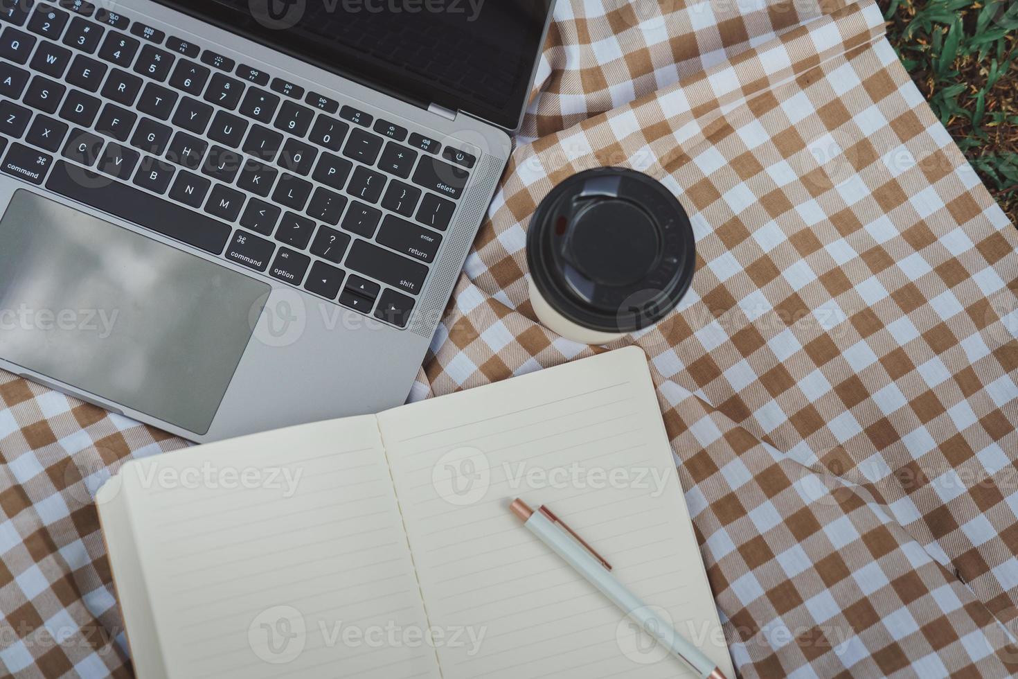 Work on laptop on picnic blanket on the lawn in the park- next to a headphone, notebook  and cup of hot coffee. Freelancer work concept. Top view, flat lay. Work form anywhere concept. photo
