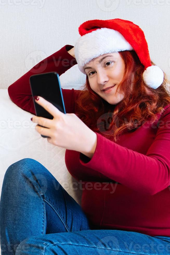 Young red-haired woman in Santa hat sits on couch at home and uses mobile phone to videochat. Online greeting with New Year and Christmas. photo