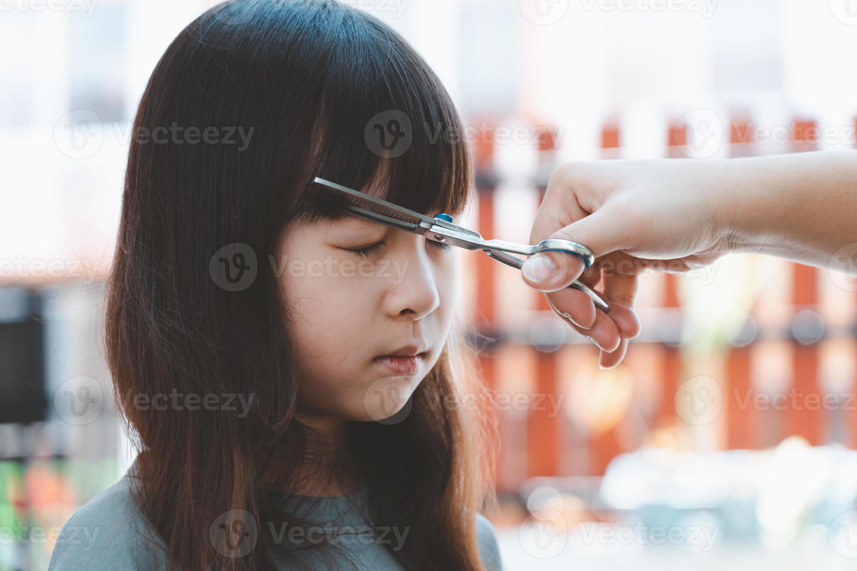 Female hand cuts hair for Asian girl with scissors and comb. Hair care