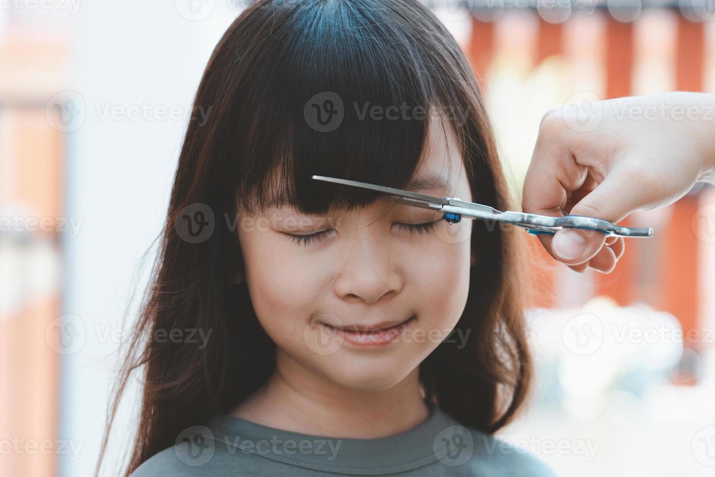 manos de mujer para cortar el pelo delantero o flequillo para una linda chica asiática en casa. las madres están felices de cortar el pelo de sus hijos. concepto de cuidado del cabello foto