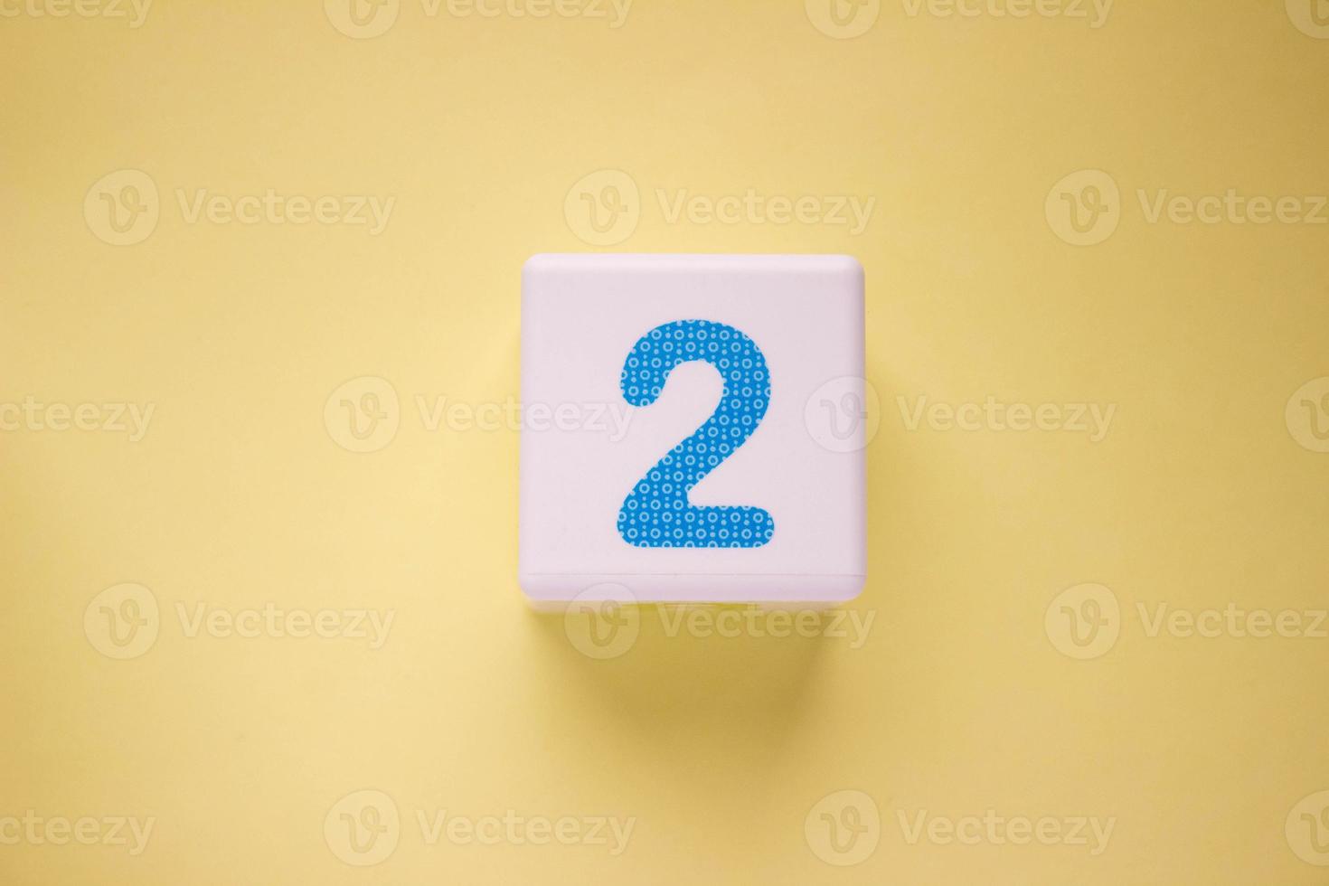 Close-up photo of a white plastic cube with a blue number 2 on a yellow background. Object in the center of the photo