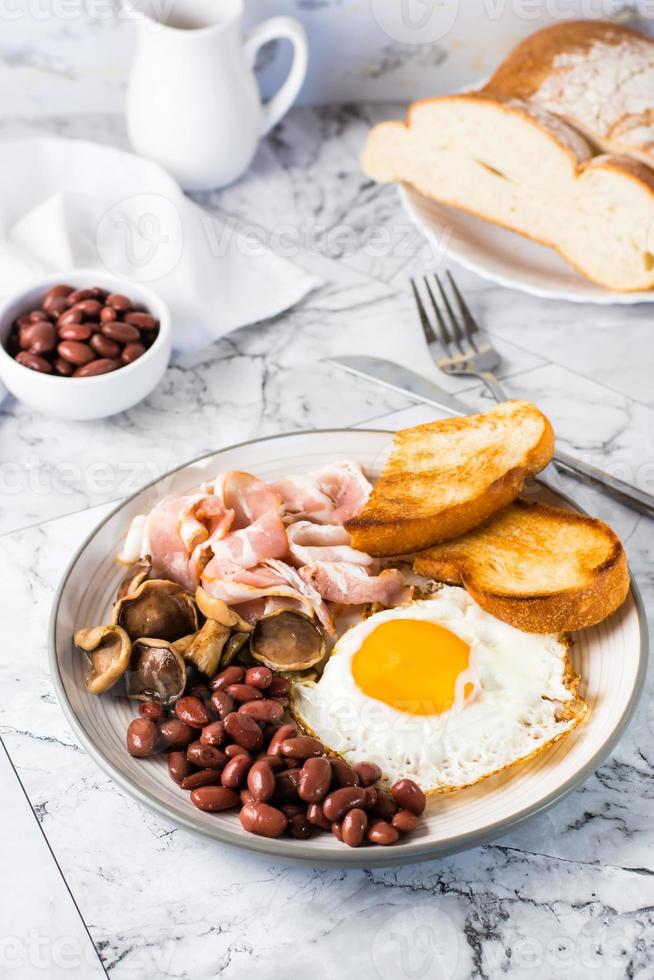 English breakfast with fried egg, bacon, beans, mushrooms and toast on a plate. Vertical view photo