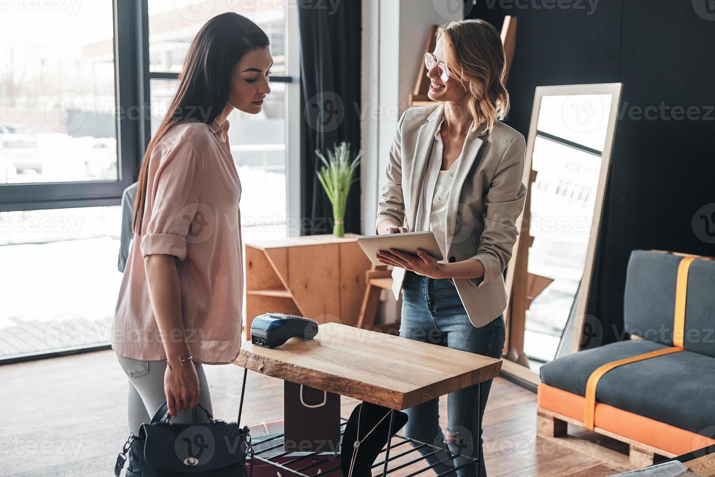 cómo le gustaría pagarle a una hermosa joven que sostiene una tableta digital y sonríe a su cliente mientras trabaja en la boutique de moda foto