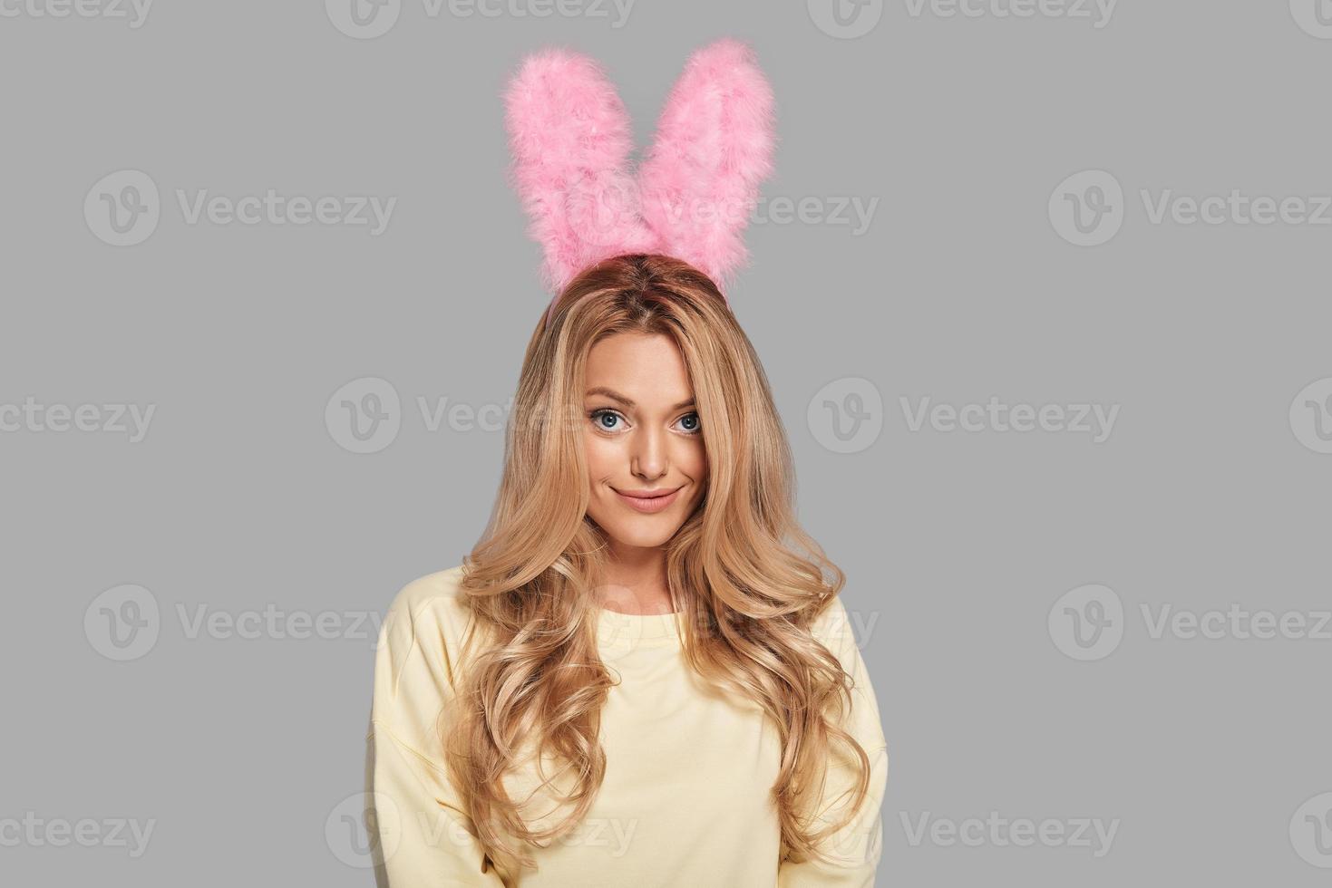 Playful beauty. Attractive young woman in pink bunny ears looking at camera and smiling while standing against grey background photo