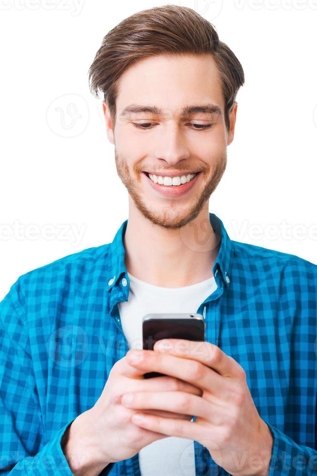My gadget is my helper. Happy young man in shirt holding mobile phone and smiling while standing against white background photo