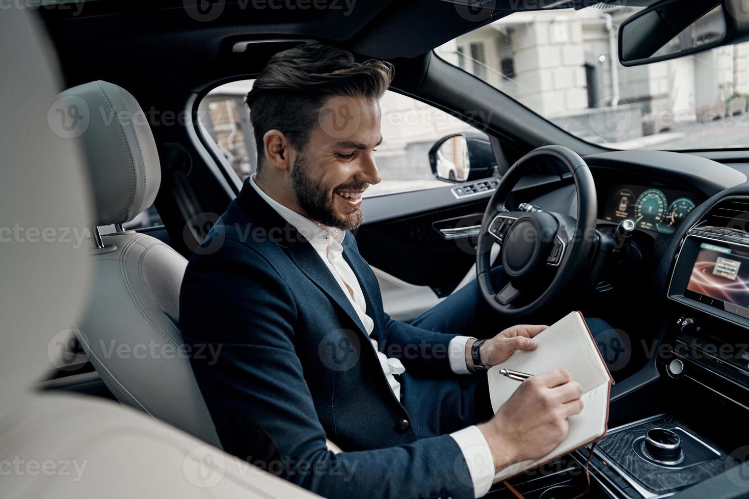 And then I am going to meet with...  Handsome young man in full suit writing something down in personal organizer and smiling while sitting inside of the car photo