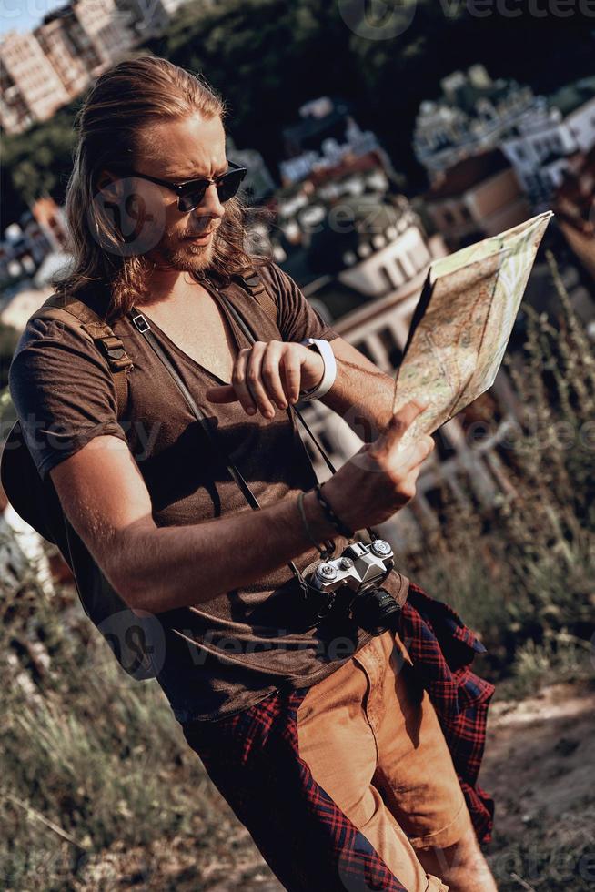 momento. un joven apuesto con ropa informal sosteniendo un mapa y revisando la hora mientras está de pie en la colina al aire libre foto