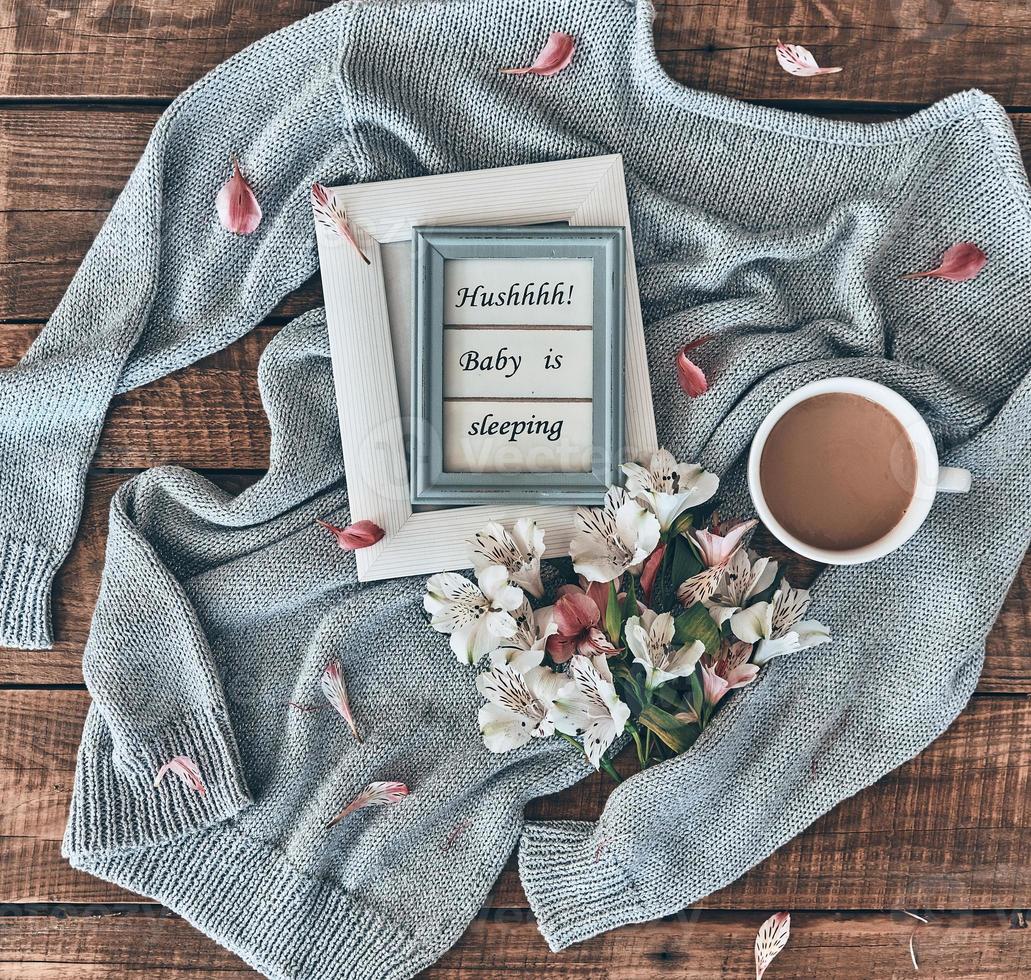Cozy concept. High angle shot of sweater, flowers, cup of coffee and photo frame on wooden desk