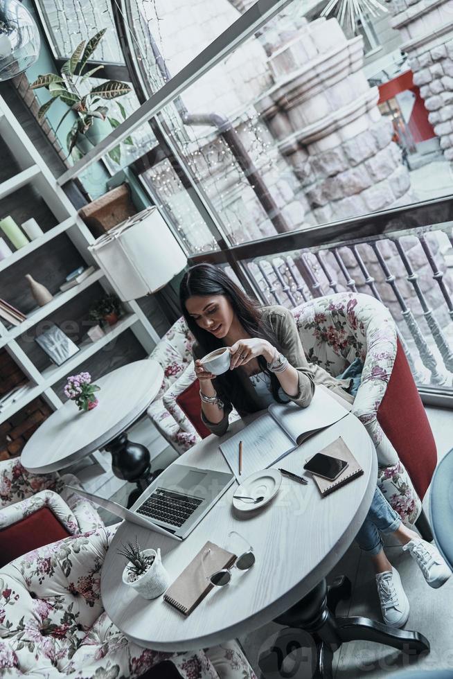 disfrutando de un café recién hecho. vista superior de una atractiva joven sonriente sosteniendo una taza de café mientras se sienta en un restaurante cerca de la ventana foto