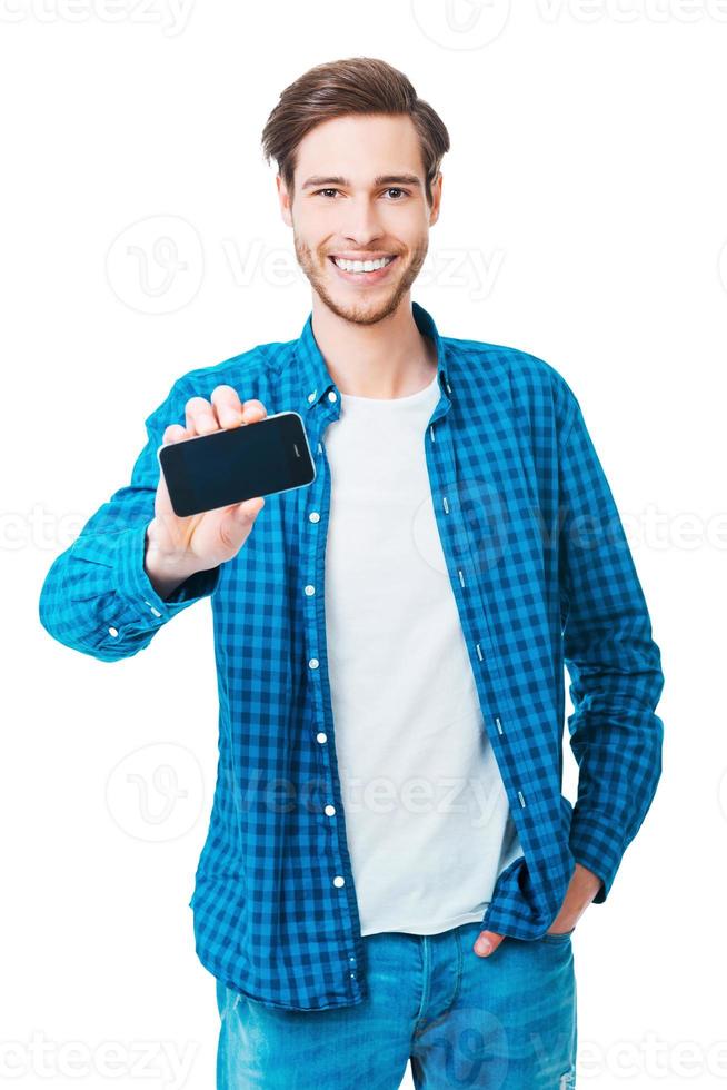 Showing his new smart phone. Cheerful young man stretching out mobile phone and smiling while standing against white background photo