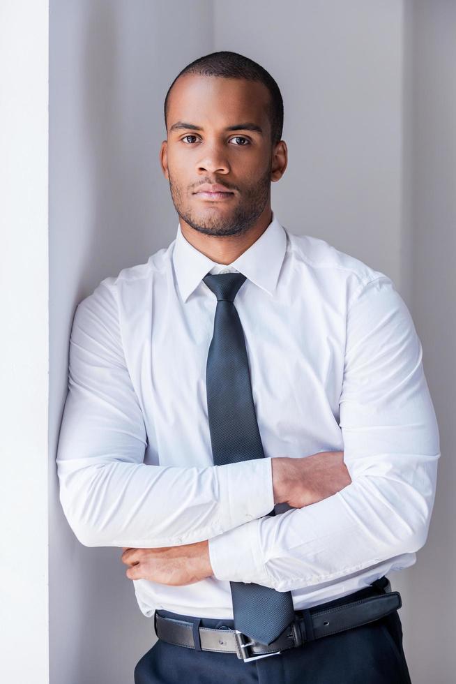 Hombre de negocios exitoso. apuesto joven africano con camisa y corbata mirando a la cámara y manteniendo los brazos cruzados mientras se apoya en la pared foto