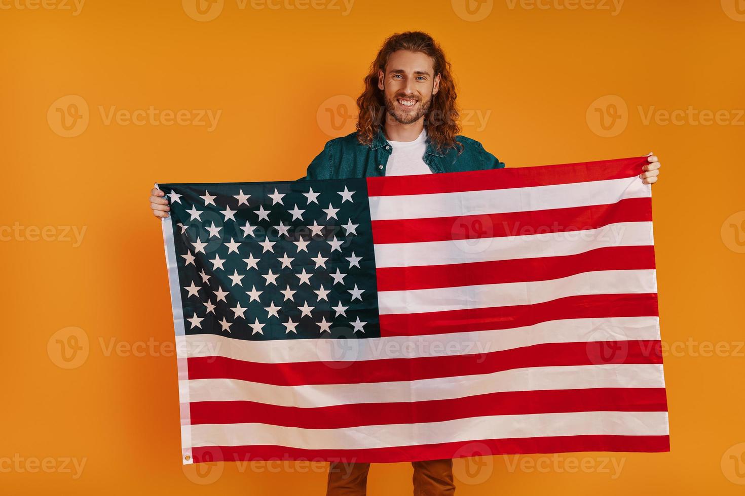 Cheerful young man in casual clothing looking at camera and smiling while holding American flag against yellow background photo