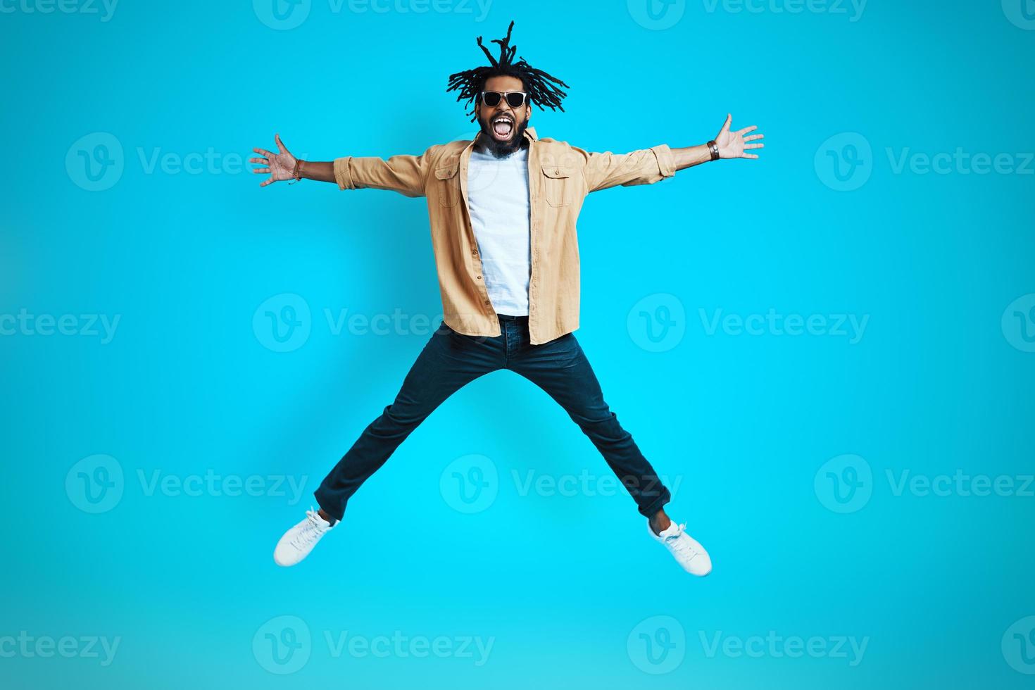 Full length of happy young African man in casual wear keeping arms and legs outstretched while hovering against blue background photo