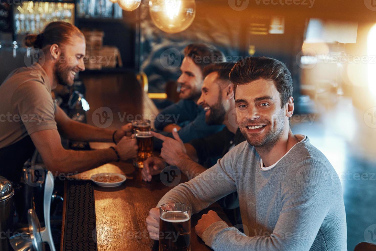 jóvenes sonrientes con ropa informal bebiendo cerveza y uniéndose mientras están sentados en el pub foto