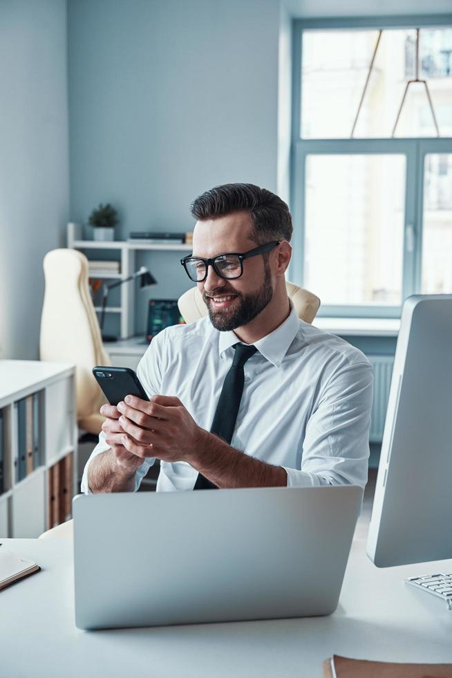 Handsome young man in formalwear using smart phone and smiling while sitting in the office photo