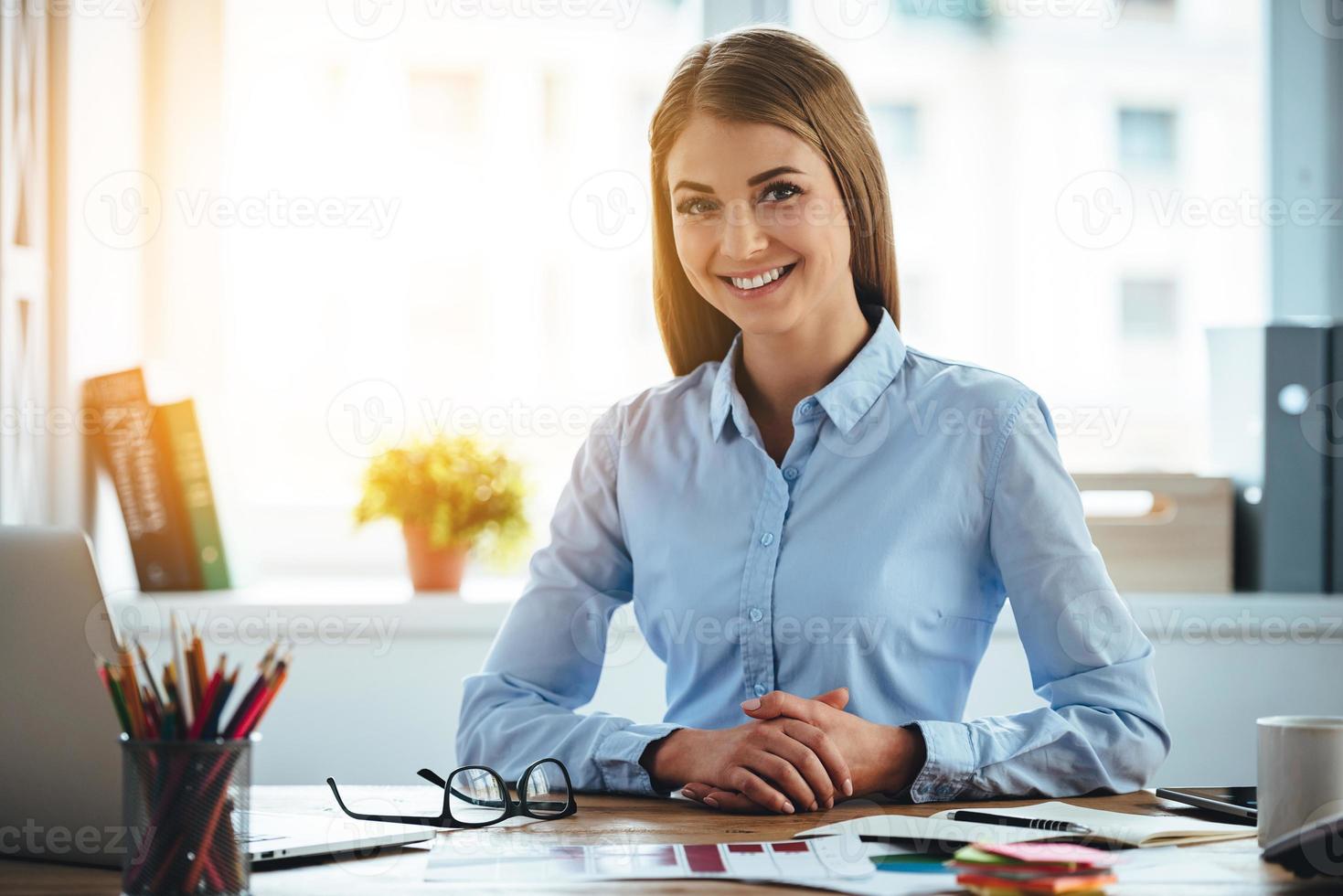 Toothy smile from real expertise. Cheerful young beautiful woman looking at camera with smile while sitting at her working place photo