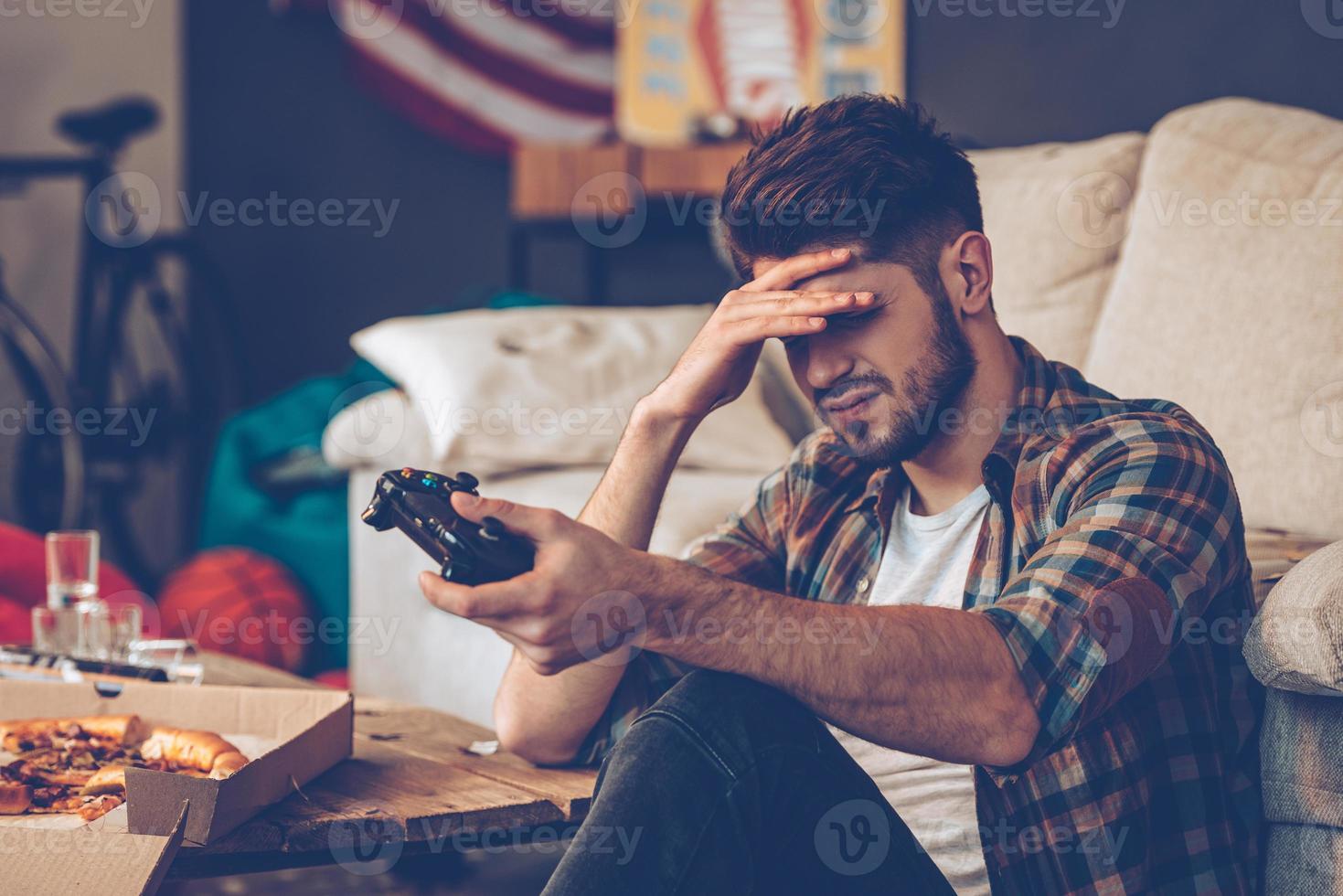 Game over. Frustrated young man holding joystick and keeping his hand on forehead while sitting on the floor in messy room after party photo