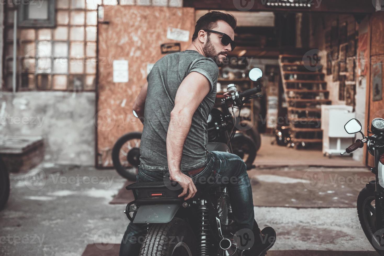 Feeling comfortable on his bike. Rear view of confident young man sitting on his bike and looking over shoulder with motorcycle garage in the background photo