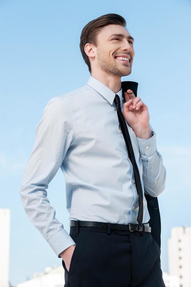 hombre de negocios feliz. apuesto joven en ropa formal sosteniendo su chaqueta en el dedo y mirando hacia otro lado mientras está de pie contra el cielo azul foto