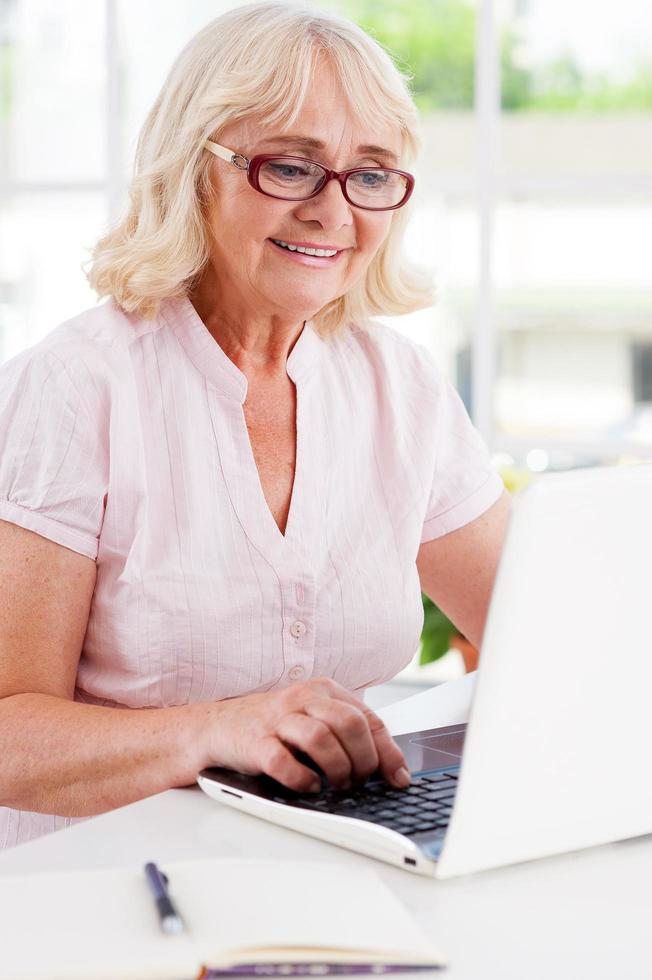 trabajando en casa. feliz anciana trabajando en la laptop y sonriendo mientras se sienta en la mesa foto