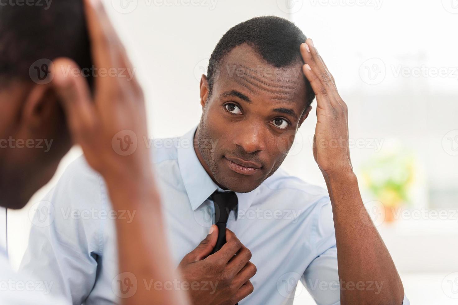 Feeling confident about his look. Young African man looking at the mirror and adjusting his necktie photo