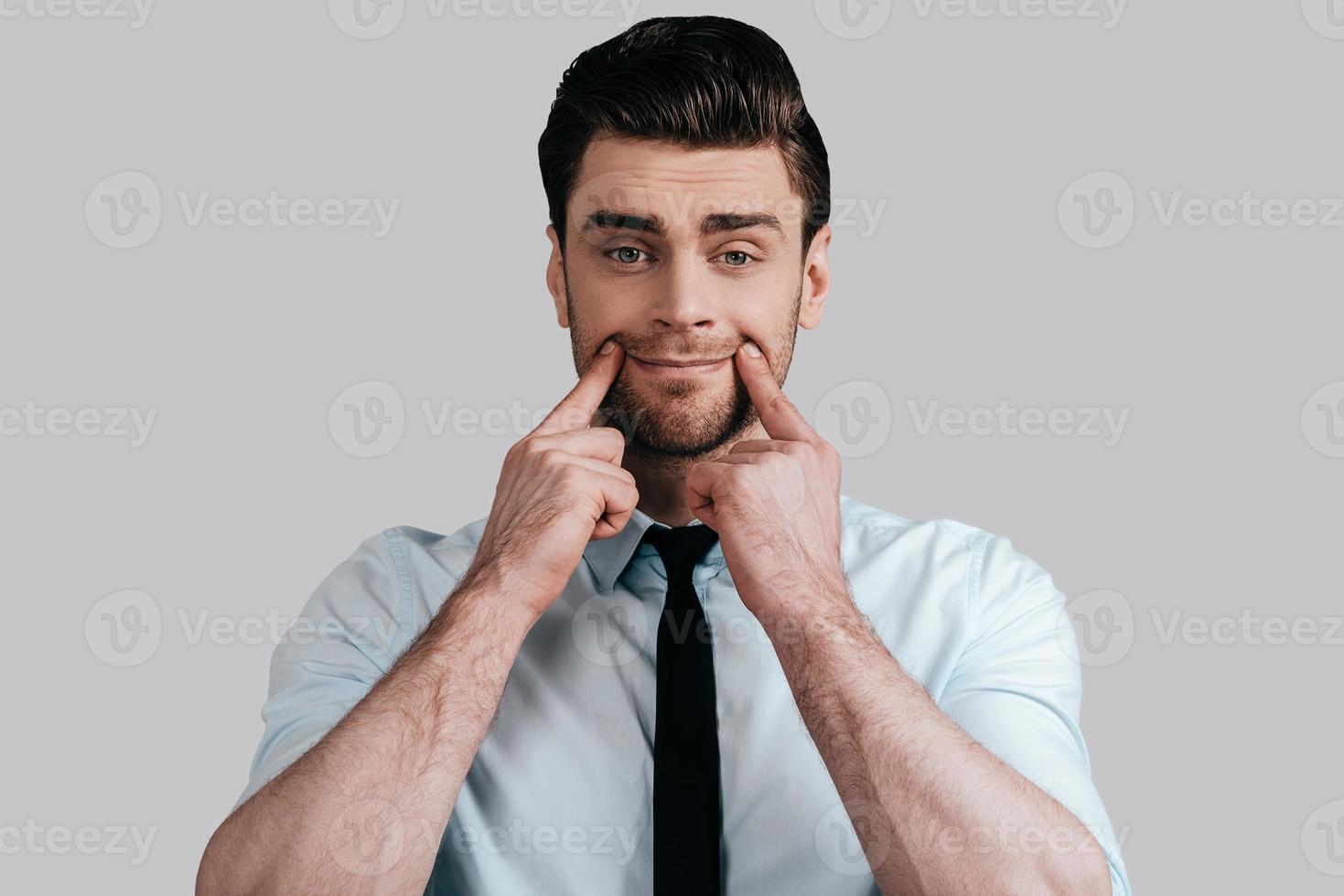 Nothing cheers him up. Sad young man in white shirt and tie trying to smile by touching his face with fingers and looking at camera while standing against grey background photo