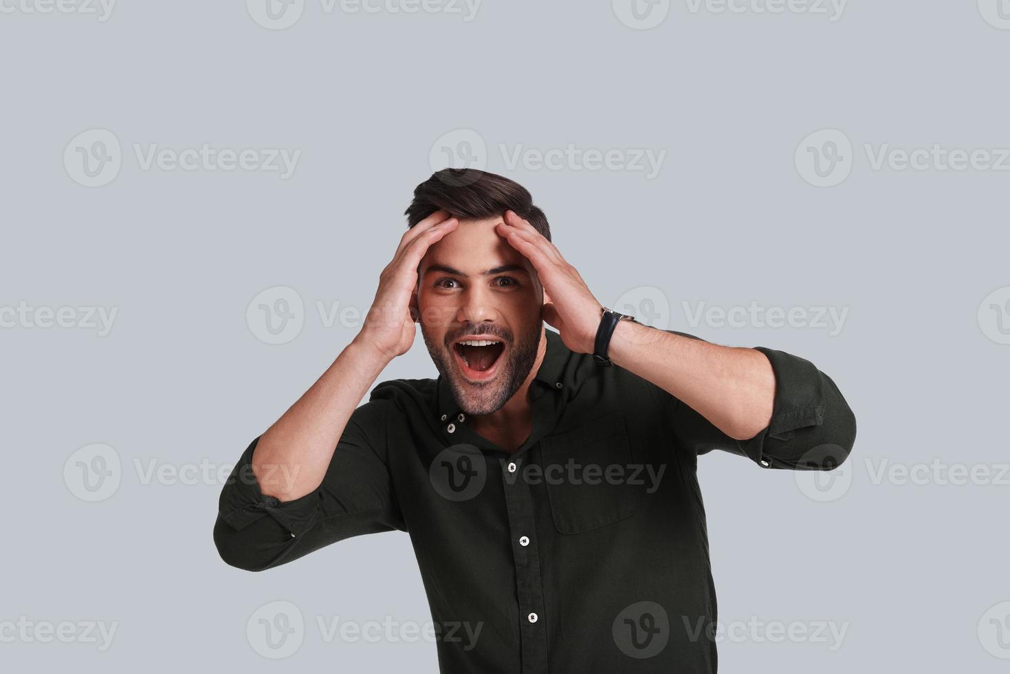 Amazing news. Shocked young man keeping mouth open and looking at camera while standing against grey background photo