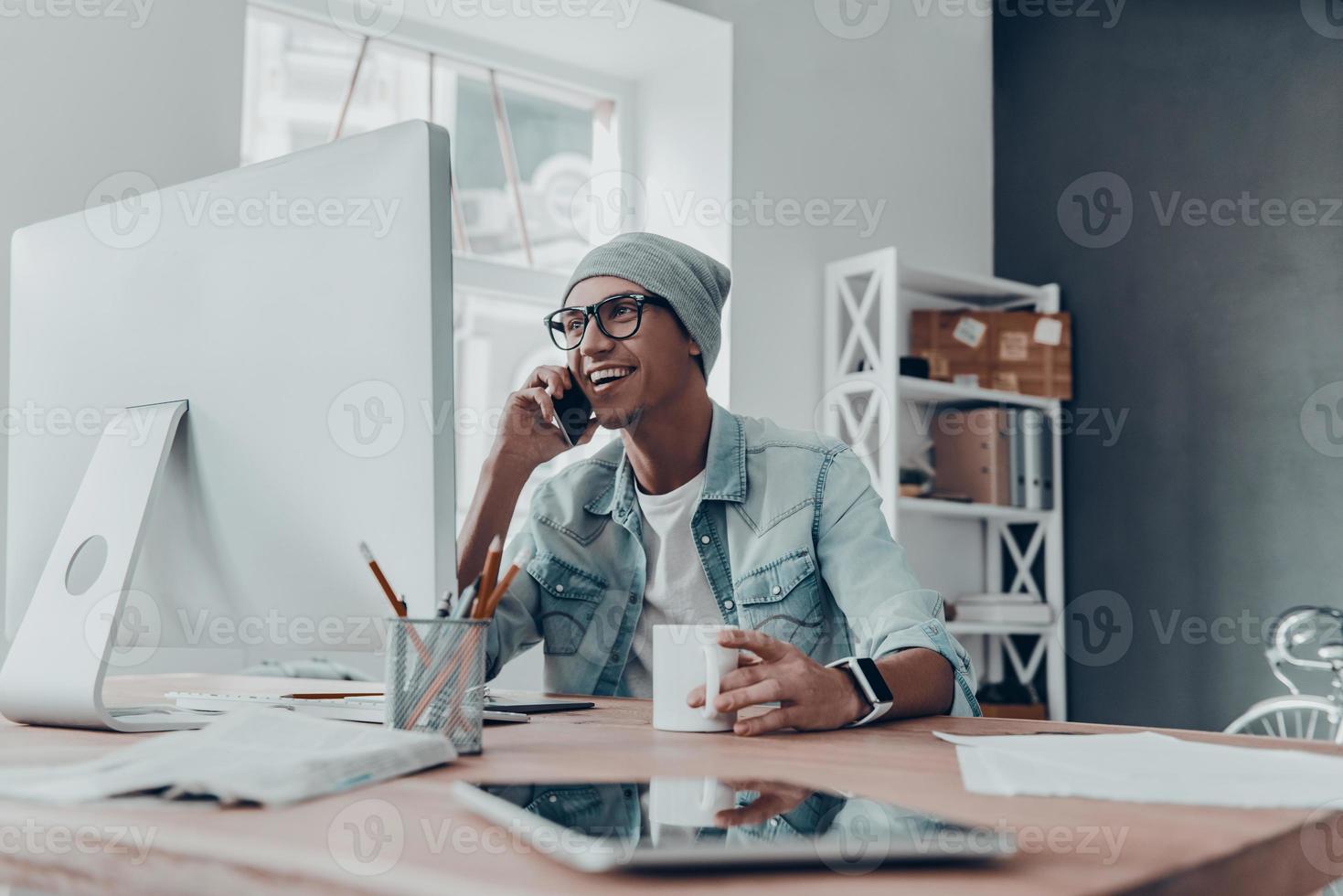 Good talk with client. Handsome young man in eyewear talking on smart phone and smiling while sitting at his working place in creative office photo