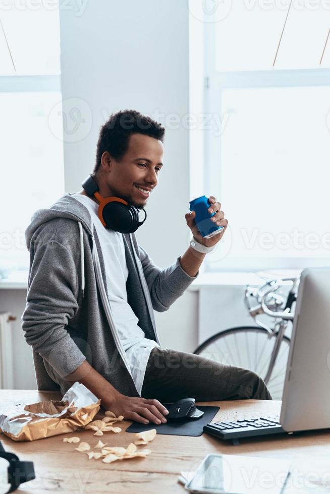 Just resting. Relaxed young man in casual clothing smiling and using computer while spending time at home photo