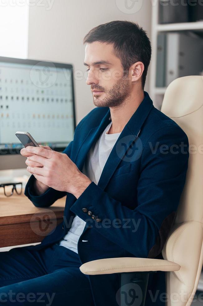Typing business message. Side view of confident young man holding smart phone and looking at it while sitting at his working place in office photo